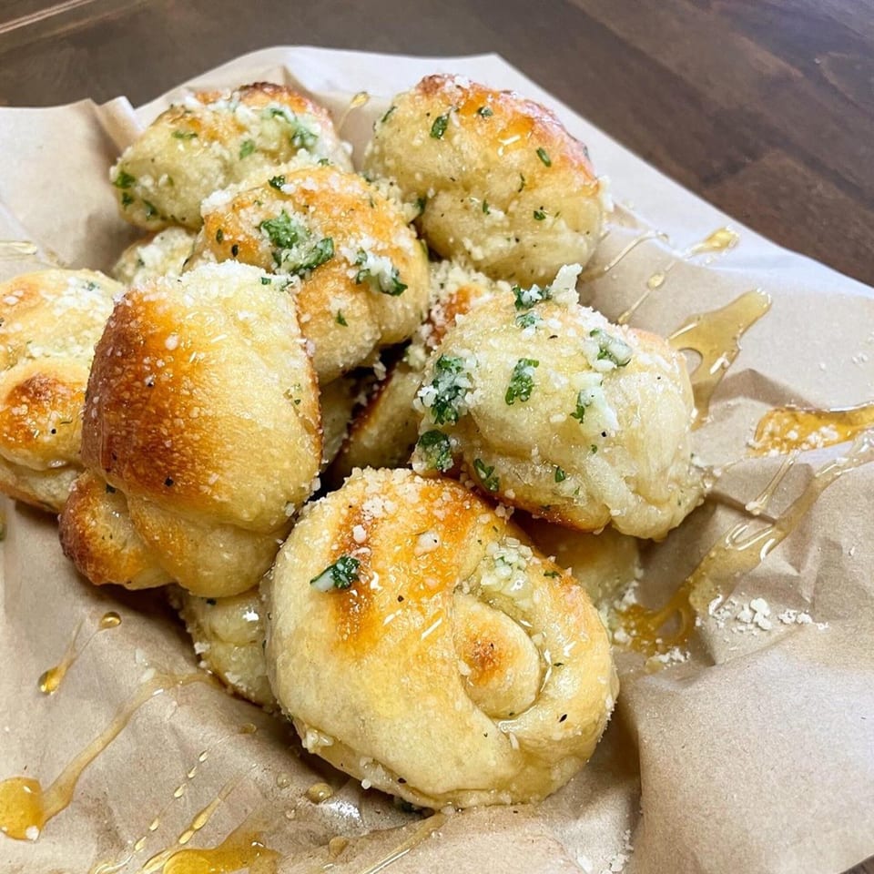 Golden-brown PEEL garlic knots with parmesan, parsley, and honey drizzle on brown paper.