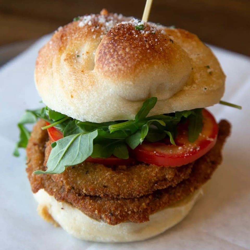 Close-up of a fried chicken sandwich on a bun with arugula, tomatoes, and parmesan cheese.